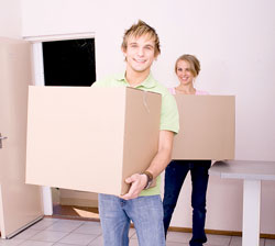 A blonde man and woman hold boxes of items they are going to donate while smiling at the camera