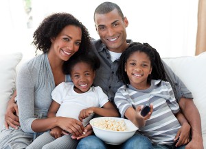 A mom and dad sit on the couch with their young daughters on their laps holding a remote and popcorn while smiling